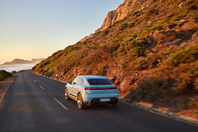 A Porsche Macan EV drives past a mountain at sunset (or maybe sunrise). The rear lights are illuminated.