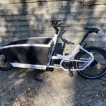 Image of a front-loading cargo bike with white metal tubes, set against stone pavement and walls.