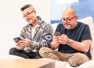 Two men both checking their smartphones as they sit together on the sofa at home.