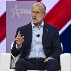 Federal Communications Commission member Brendan Carr sits on a stage and speaks while gesturing with his hand. Behind him is the CPAC logo for the Conservative Political Action Conference.