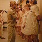 Blond man dressed as prison guard wearing sunglasses, harassing prisoners in hospital gowns.