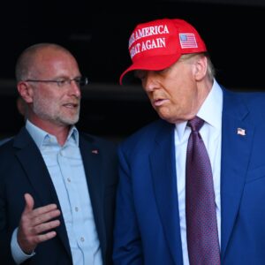 FCC Commissioner Brendan Carr standing next to and speaking to Donald Trump, who is wearing a "Make America Great Again" hat.