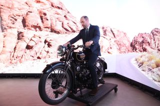 Prince William riding a motorcycle in a suit in front of a green screen with a desert road