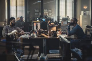 Isaiah Mustafa as John Sampson and Aldis Hodge as Alex Cross, facing each other as they sit at desks in a police station, in 'Cross.'