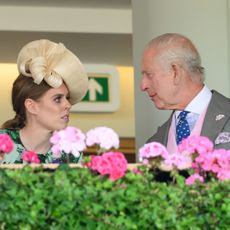 Princess Beatrice wearing a straw hat and green dress speaking with King Charles in a gray suit at Royal Ascot with pink flowers in the foreground