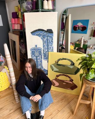 Ella Emhoff sitting on the floor in front of two knit paintings of blue cowboy boots and two handbags