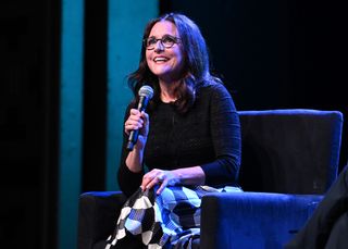 julia louis dreyfus wearing glasses and a patterned skirt on stage at a wiser than me podcast event