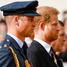 Prince William, wearing a blue military uniform, and Prince Harry in a suit and tie standing next to each other looking solemn at Queen Elizabeth's funeral