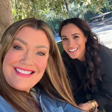 Jamie Kern Lima and Meghan Markle smiling next to each other in a selfie outside with a tree behind them