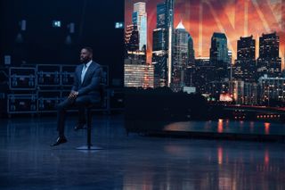 Colman Domingo as Muncie Daniels, sitting on a stool in an empty set, with a backdrop of a cityline sunset behind him, in Episode 101 of 'The Madness.'