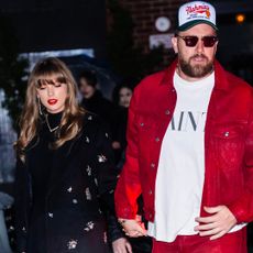 Travis Kelce wearing a red jacket and white tee walking next to Taylor Swift in a black coat walking into a restaurant