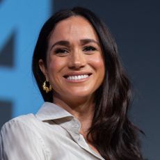 A headshot of Meghan Markle wearing a tan blouse and gold hoop earrings smiling in front of a blue background 