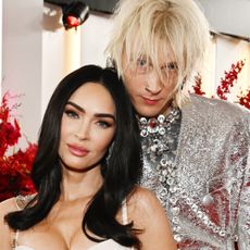 Megan Fox is smiling and has long black hair while Machine Gun Kelly stands behind her with shaggy blonde hair and wearing a silver suit