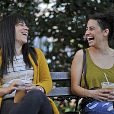 abbi and ilana sit on a park bench laughing in broad city