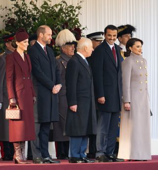 Kate Middleton wearing a marroon coat, hat and boots standing next to Prince William, King Charles, the Amir of Qatar and Sheika of Qatar