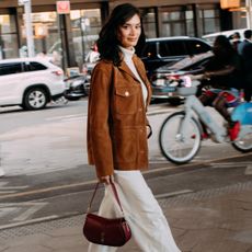 a guest attends new york spring summer 2025 fashion week in a brown suede jacket, white pants, carrying brown bag