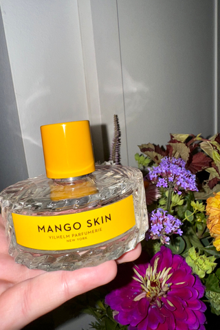 A hand holding Vilhelm Parfumerie Mango Skin Eau de Parfum in front of flowers