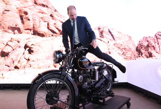 Prince William climbing on a motorcycle in a suit with a desert road virtual background behind him