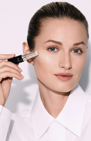 Woman in a white shirt holding Cle de Peau concealer stick.