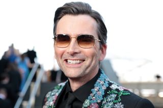 David Tennant wears sunglasses and a black suit embellished with floral design on a red carpet