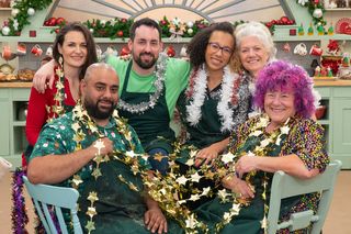 several great british baking show holiday contestants standing around a table and chairs with star decorations and tinsel