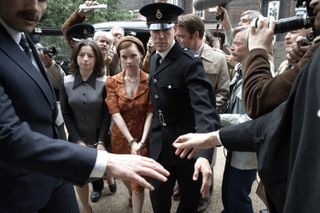 Hazel Doupe as Marian Price (left, wearing black) and Lola Petticrew as Dolours Price (left center, wearing orange) are led handcuffed through a crowd with policeman ahead of them and reporters behind them, in 'Say Nothing.'