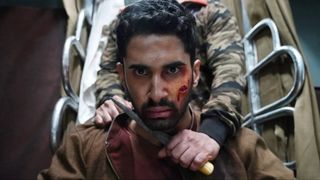 A close-up on a man's (Lakshya) bloody face, as an attacker holds a knife to his throat while standing in front of silver bars, in 'Kill.'