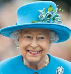 Queen Elizabeth wearing a blue hat with flowers on it and a matching blue coat with pearls and smiling