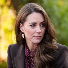 Kate Middleton wears a brown blazer with a burgundy and white polka dot blouse, and has long wavy brown hair, as she looks at Prince William, who is wearing a suit and tie and has beard stubble