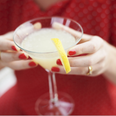 Woman in red with red nail polish holding alcoholic drink with a slice of lemon garnished on the rim of the glass