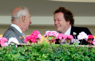 King Charles and Tom Parker Bowles wearing suits and smiling while talking to each other in front of a row of pink flowers