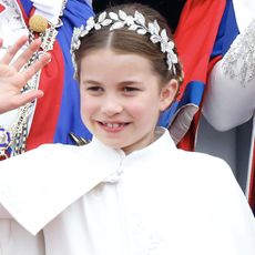 Princess Charlotte wears a white dress with a white cape and a floral head band as she waves and Queen Elizabeth wears pearls and a green paisley dress