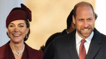 Kate Middleton wearing a maroon coat and hat and smiling next to Prince William wearing a dark coat and a red tie 