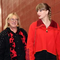 Taylor Swift wearing a red pullover walking next to Donna Kelce next to a red brick wall