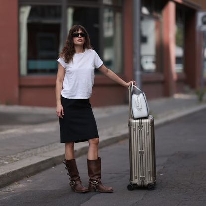 Lea Naumann is seen wearing black sunglasses from Prada, a white tee from Mother Denim, a black knee-length vintage skirt, brown leather boots with silver buckle closure from Miu Miu, a white leather Supernova bag from Prada and a silver suitcase from Rimowa on July 11, 2023 in Berlin, Germany.