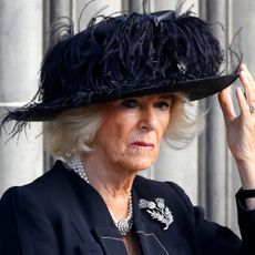 Queen Camilla wears a feathery black hat and looks sad