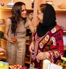 Meghan Markle holding a bowl and putting her arm around a woman wearing a black head scarf and colorful red outfit