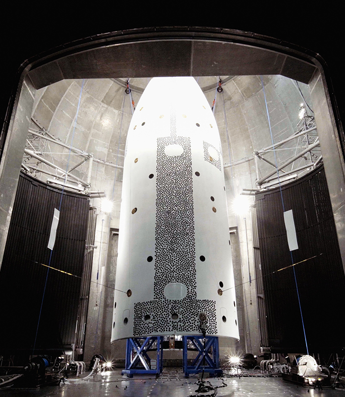 A tall, white cylindrical structure with a cone-shaped nose stands inside a grey testing chamber on a blue metal scaffolding structure.