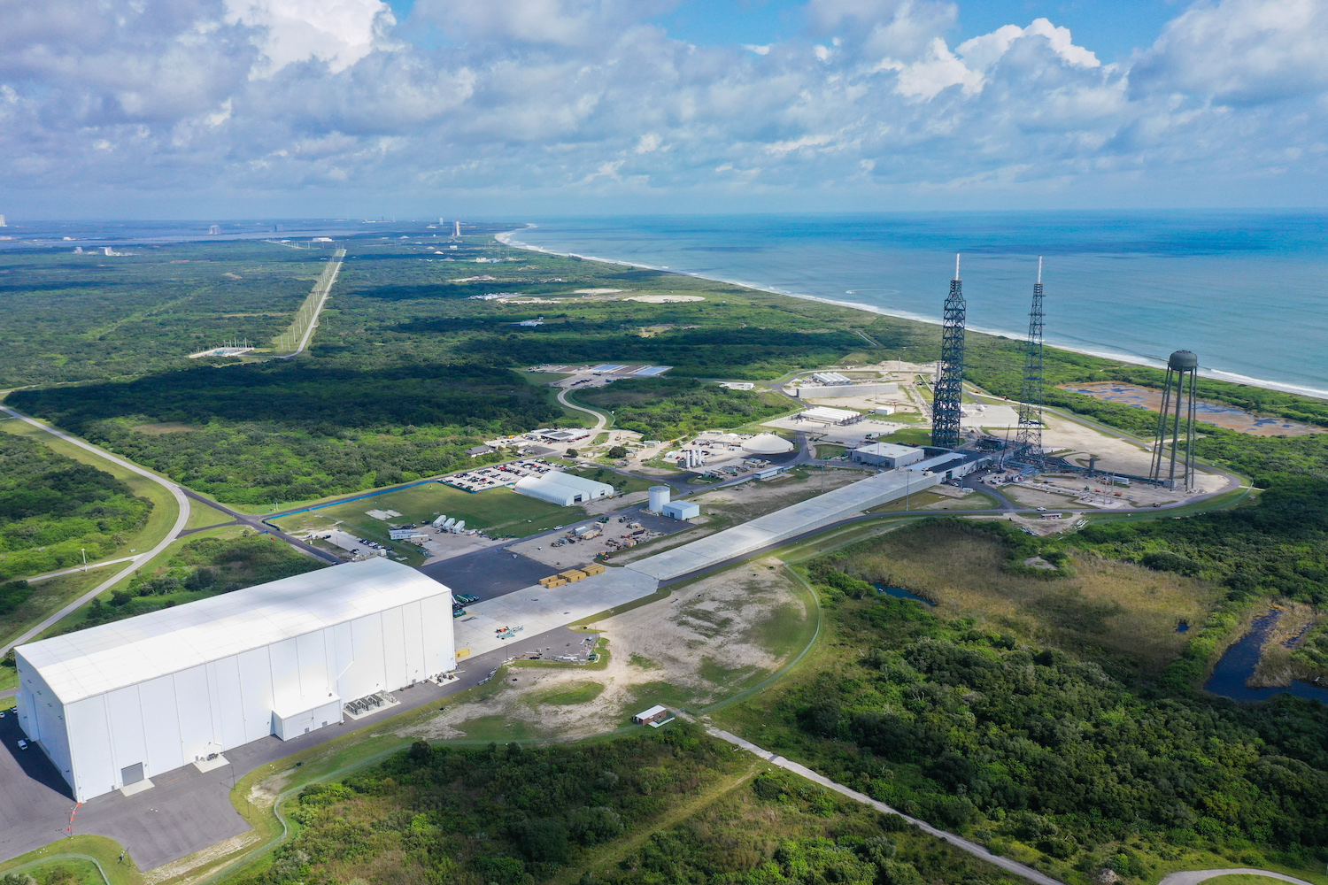 An aerial view of Launch Complex 36 shows the Cape Canaveral coastline. One of Blue Origin’s large white buildings is visible in the foreground; lightning towers and the water tower are in the distance.