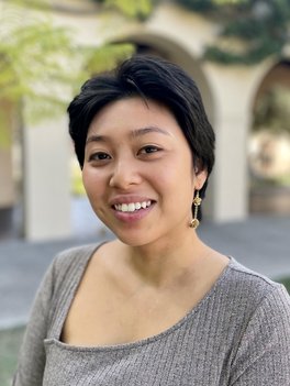 An image of a young Asian woman with short dark hair smiling, wearing a light grey sweater top with an asymmetrical neckline