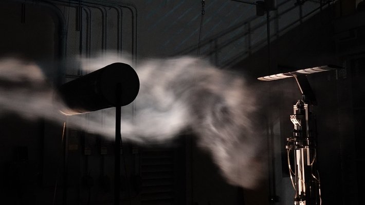 The turbulent wake of a cylinder can be seen in illuminated smoke in a darkened wind tunnel. An airfoil instrument is set up on the right side of the image. A large cylinder is on the left side of the image, creating turbulence.