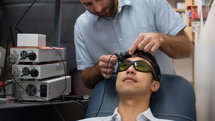 One man is putting a headset-based device on another man who is sitting in a chair in front of him.  The man in the chair is wearing sunglasses