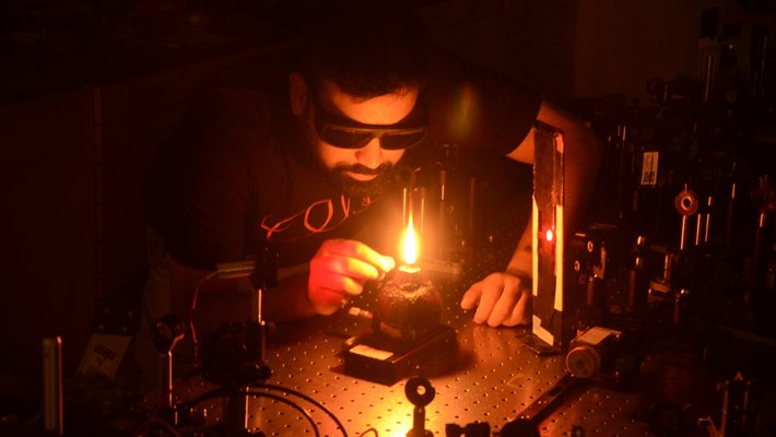 A man with a beard, wearing goggles holds a device while looking into a flame within an optical instrument setup in a lab.