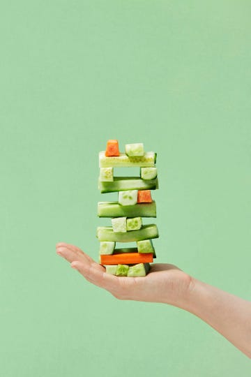 a hand supports a vertical arrangement of colorful vegetable pieces primarily composed of cucumbers and carrots neatly layered in a playful, balanced fashion the background is a soft green complementing the fresh tones of the vegetables