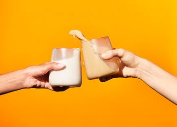 two hands holding milk based drinks clinking glasses in front of an orange background