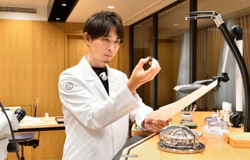 a person in a white lab coat holding a small object in one hand and a piece of paper in the other with a workbench and tools visible in the background