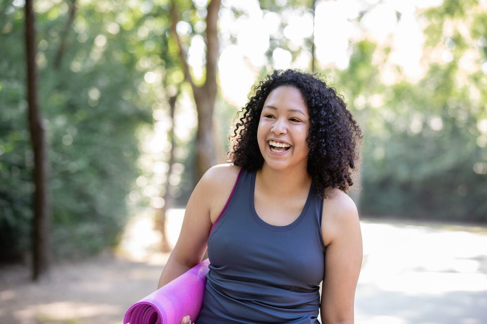 woman smiles while holding yoga mat and walking outdoors for exercise