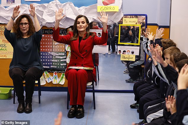 Letizia sits in a classroom with children and interacts with them by putting her hands up