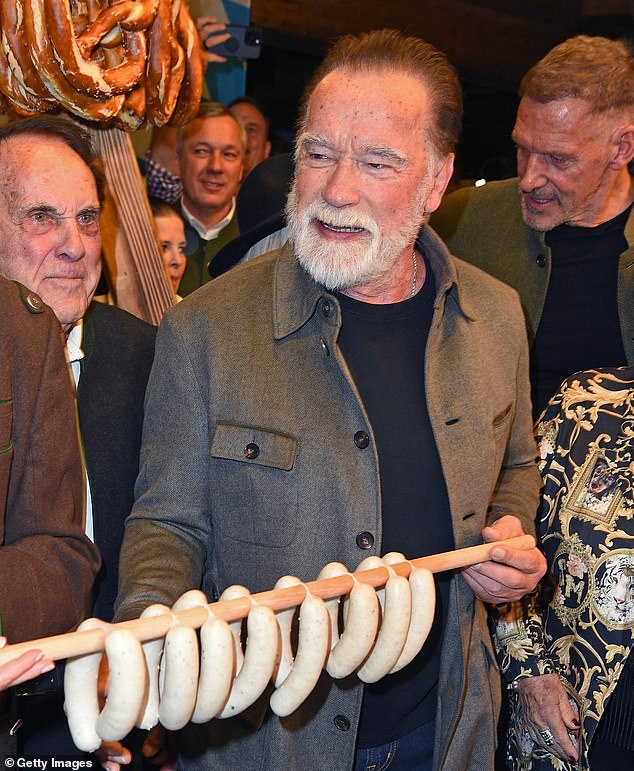 Arnold wore a grey jacket over a navy sweater, adding some mid blue jeans and a silver necklace, as he displayed the Bavarian sausages which were hung over a long wooden spoon