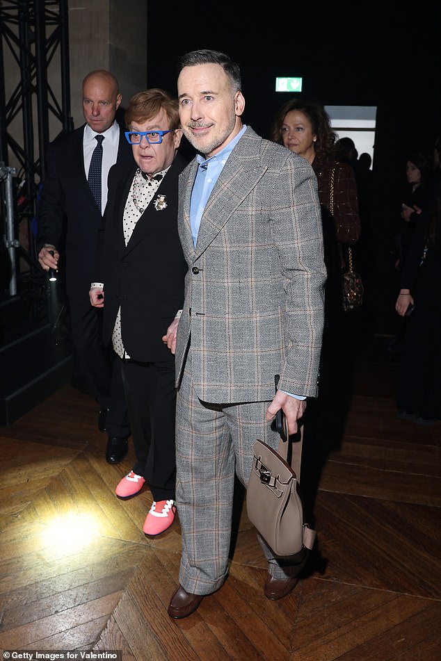 He accessorised the look with white and pink pearl necklace and bright red trainers as he sat beside his husband David Furnish